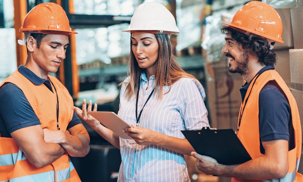 A woman is speaking to two men; all wearing hard hats. The two men are dressed in orange hi-vis vests and are listening attentively.