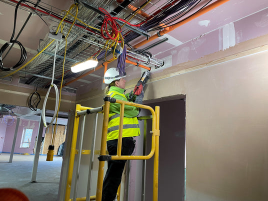 Image of a woman wearing a hard hat and high viz jacket, using the Quelfire intumescent sealant to seal around a pipe through a wall. There are many exposed wires and cables running across, and hanging from, the ceiling.