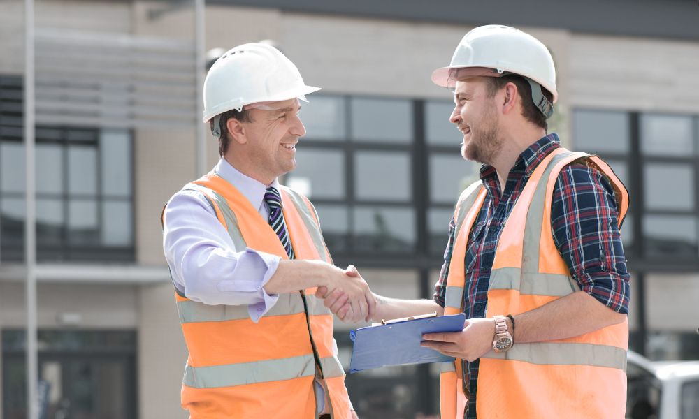 Two men wearing orange high-visibility vests smiling and shaking hands in front of a building.