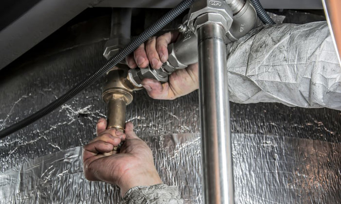 Close-up of hands working on metal plumbing pipes beneath a surface, wearing white protective gear.