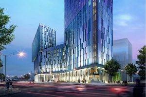 A modern, glass-clad building at dusk with illuminated interiors. The structure has dynamic geometric shapes, trees lining the street, and a peaceful, futuristic ambiance.