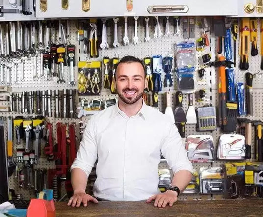 Smiling man in a white shirt stands in front of a wall of tools, at a wooden counter.