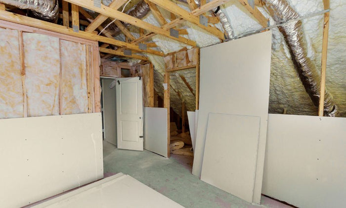An unfinished attic space with exposed wooden beams and insulation. Drywall sheets are propped up against the walls and there is an open door. 