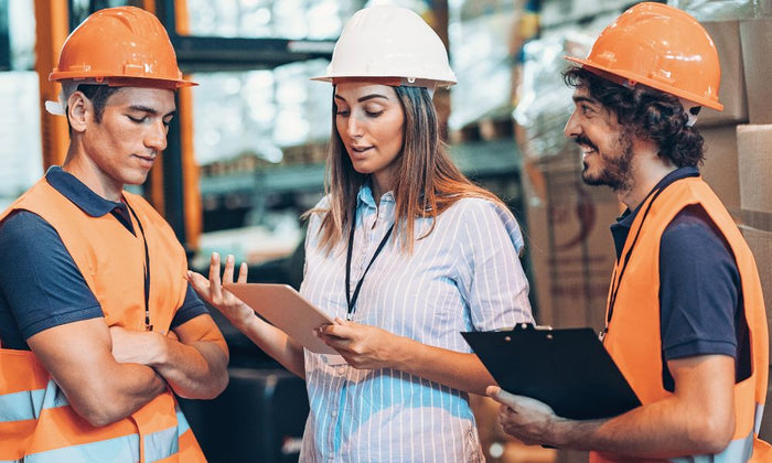 A woman is speaking to two men; all wearing hard hats. The two men are dressed in orange hi-vis vests and are listening attentively. 