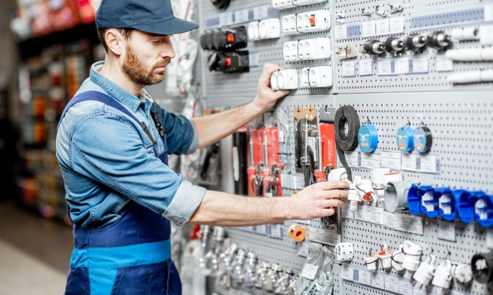 A male with a beard in a blue shirt, blue apron and cap examining or organising electrical components such as sockets and plugs, on a pegboard in a hardware store.