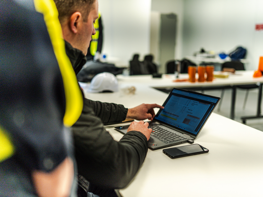 Sales Director, Craig Wells, wearing a black jacket, pointing to a laptop screen. In the background, you can see three tubes of Quelfire biscuits and a white hard hat. 