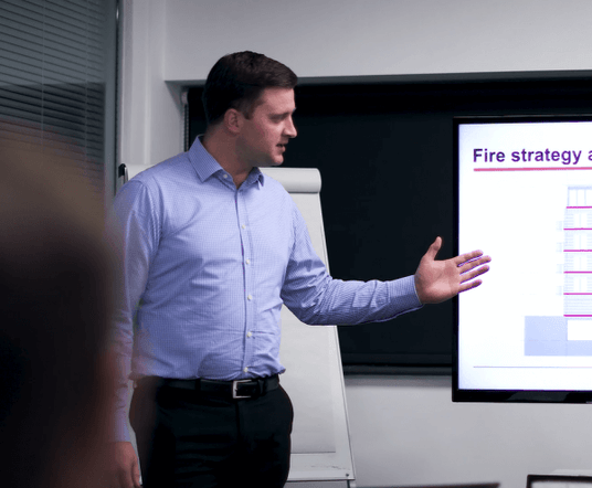 Image of Craig Wells, Sales Director, wearing a blue shirt, presenting. He has a flip chart behind him and he is pointing to a large presenter screen, which we are unable to see much of.