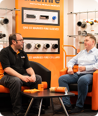 Alec (technical) and Glenn (technical) are sat with hot drinks and biscuits in the training room. A demo board with Quelfire products can be seen behind them.