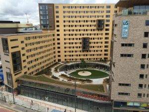 A large, modern U-shaped building surrounds a circular courtyard with landscaping and a central circular feature. The sky is cloudy.