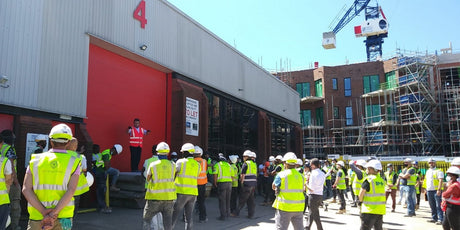 Construction site is displayed with a guy at the front wearing a pink high viz jacket, talking to a crowd of other men in yellow high viz jackets and hard hats. 